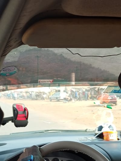 The image captures a first-person perspective from inside a car, looking out through the windshield onto a bustling roadside scene. The foreground shows parts of the car's interior, including the steering wheel on the bottom right, with a hand visible on it, suggesting someone is driving or stationary. A red and black phone holder is mounted on the left side of the dashboard. On the right side of the dashboard, there's a small soccer ball ornament, a clear plastic bottle, and an orange container with an unreadable label. A visible crack runs across the bottom of the windshield.
Outside the car, a dusty, unpaved area functions as a market or informal gathering space. Numerous people are present, clustered around various stalls or small structures. Several vehicles, including a light-colored car and an orange/red sedan, are parked or moving slowly through the crowd. In the background, what appears to be a distant sign bears the text "Oando," a Nigerian petroleum company. Further behind, hazy, mountainous terrain dominates the horizon under what appears to be a bright, possibly dusty, daytime sky. On the top left of the windshield, a sticker is visible with "SOSO" and other unreadable text. The overall scene suggests a vibrant, semi-urban or rural setting, likely during the day, characterized by a lively community atmosphere.