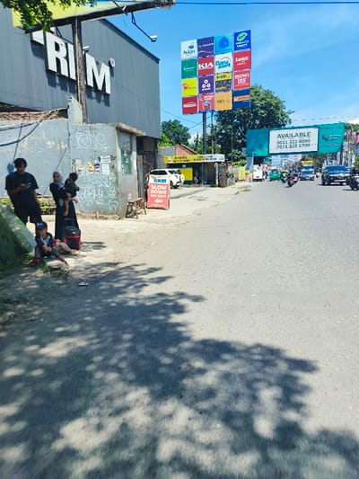 The scene takes place on a street in Kecamatan Margahayu, Indonesia, with a large building displaying the word "RIM" on the side. Three individuals stand on the sidewalk, two adults and one child.  The adult female is holding a young child and both are wearing colorful clothing. The adult male is standing to the left and holding a cell phone. A child stands to the left of the adult female, wearing a black and white shirt and blue jeans. The child is looking down at something on the ground. A large billboard with a variety of colorful advertisements is located on the opposite side of the street. The street is mostly empty, except for a few vehicles and a person crossing the road. The street has a gray asphalt surface. Trees line the street on both sides. The sky is a clear blue, indicating a sunny day.  There is a shadow cast across the street from a tree, suggesting a large tree is off camera.  A red sign with white letters on it is next to a white car, with the words "Slow Down" in a square box, and the words "Rim Kodo" underneath.
