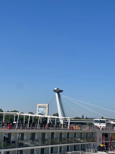 The image depicts a sunny day in Bratislava, Slovakia, with the iconic UFO Observation Deck atop the New Bridge (Most SNP) prominently featured against a clear blue sky. In the foreground, a white river cruise ship, identified by lettering reading "BRATISLAVA SK", is docked. The ship has multiple decks with cabins visible, featuring glass railings and doors. The upper deck of the ship shows outdoor seating areas, some with umbrellas, and bicycles. A red cylindrical object and an orange life preserver are visible on the upper deck. Further in the background, behind the cruise ship and a line of trees, a tall building with a sign that reads "INCHEBA" is visible. Several people are visible on the ship, including two individuals on the lower deck, one wearing a yellow shirt and the other a dark blue shirt, who appear to be crew members. The overall scene conveys a sense of urban riverfront activity on a bright day.