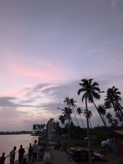 The image depicts a beachside scene in Badem, India, during twilight. The sky is a gradient of pale purple and pink, with wispy clouds. A calm body of water is on the left, bordering a paved walkway lined with palm trees. Several people are visible on the walkway and on the beach. On the walkway, a group of individuals is gathered, some looking out at the water, while others are seated. A red and white banner displaying the word "KAYAKING" is planted near the walkway. A few cars are parked in the background, near a dense line of palm trees. The scene suggests a relaxed atmosphere, possibly at the end of the day, with recreational activities like kayaking being offered.