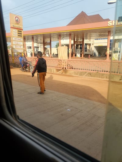 The image shows a street scene in Kakau, Nigeria, featuring a SHARON gas station. The gas station has a prominent sign with the brand name and categories like Petrol, Diesel, Kerosene, Lubricants, Autocare, Shop, and Milk & Snacks. Several fuel pumps are visible under the covered area of the station.
In the foreground, a man wearing a black hoodie and orange pants with black stripes is standing facing away from the camera. His posture suggests he is either waiting or observing something. To his left, on a motorcycle, are two men. The man in the front is wearing a blue shirt, and the man behind him is wearing a checkered shirt. They appear to be interacting or waiting.
The ground is a mix of paved concrete and dirt. The overall environment suggests an outdoor, possibly commercial area. The lighting indicates it's daytime, with shadows cast by the structures and people. The view appears to be from inside a vehicle, as evidenced by the blurred foreground elements suggesting a window.