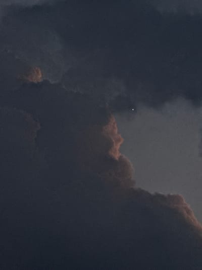 The image depicts a cloudy sky, possibly during twilight or a storm, with a single, bright celestial object visible in the upper left quadrant. The clouds are dark and dense, with some appearing to have a reddish hue, suggesting they are illuminated from behind or by the setting/rising sun. The overall mood is dramatic and somewhat ominous due to the dark clouds. The celestial object appears as a small, intensely bright point of light, possibly a planet or a very bright star. There is no text visible in the image.