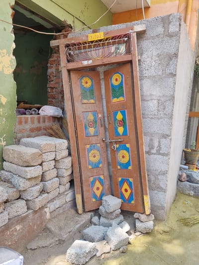 An outdoor scene in Pedda Kammavaripalli, India, depicts a construction or renovation site. A detached brown wooden double door frame, featuring an arched wrought-iron grille with yellow, red, and orange concentric rings, is propped upright. The door's panels are adorned with colorful, faded, painted designs in yellow, red, blue, and green, depicting flowers and geometric shapes. A silver-colored metal latch mechanism is visible on the right door panel. The door rests on several rough gray concrete blocks on the ground, leaning against two distinct wall structures.
To the left, a section of wall painted faded green-yellow exhibits significant peeling, revealing reddish-brown brickwork. An open doorway or large aperture in this wall exposes an interior cavity with exposed brick. On a narrow ledge within this opening, three rolls of material (one purple, two white with patterns) and a white plastic bag are visible. Below this, a stack of approximately 14-16 rectangular gray concrete blocks is neatly arranged. A natural fiber broom leans against the wall nearby.
To the right of the door, a wall constructed from gray concrete blocks, some textured and some with a smoother light gray finish, is visible. At the top of the door frame, a yellow sign with the inscription "2-322" is affixed. The ground in the foreground consists of dirt, with patches of fine yellow powder, possibly construction dust or cultural powder. Scattered small concrete fragments lie near the base of the door. Further to the right, a dark brown pot with yellow contents, possibly flowers or petals, sits on an elevated surface. No individuals are present in the scene. The area is brightly lit by daylight.