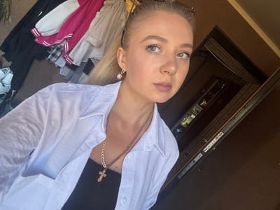 A young Caucasian woman with blonde hair tied back and light makeup is pictured in a selfie taken indoors. She is wearing a white open-collared shirt over a black tank top and a rose gold chain with a cross pendant. A rose gold earring with a flower design is visible on her left ear. In the background, various items of clothing are visible, including black, white, and pink garments, and a glimpse of a doorway or interior hallway. The scene is likely a domestic setting within Makiivka, Ukraine.