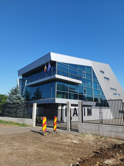 The image shows a modern, multi-story building with a glass facade and a silver-colored exterior. The building is located in Bucharest, Romania, and is identifiable by the presence of the Romanian flag and the European Union flag displayed on a balcony. The building appears to be an office or administrative complex. In front of the building, there is a black metal fence with a concrete base. Two tall traffic cones with red and yellow stripes are visible in front of the fence, suggesting ongoing construction or road work. The ground in front of the fence is covered in dirt and loose soil, indicating that the area is under development or renovation. A large evergreen tree is planted to the left of the building, partially obscured by the fence. The sky above is a clear, bright blue, suggesting a sunny day. The overall scene depicts a contemporary architectural structure within a developing urban environment.