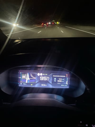 The image presents a nighttime view from the driver's perspective inside a vehicle on a multi-lane highway in Illkirch-Graffenstaden, France. The road ahead is dark, illuminated primarily by vehicle lights. White lane markings are visible on the asphalt. Multiple vehicles are observed ahead, indicated by their red rear lights and a bright white light from an approaching or distant vehicle on the left. Further down the highway, several yellow or orange lights are present, suggesting additional vehicles or road features. The vehicle's windshield displays some reflections and minor streaks.
The foreground is dominated by the vehicle's digital instrument cluster and a portion of the dark-colored dashboard. The instrument cluster centrally displays the current speed as 95 km/h, with "D" and "km/h" below it. To the left, a navigation graphic shows a blue road segment with white lines and a directional indicator. Below this, "745 km" is visible, likely indicating remaining distance. The odometer displays "19748 km". On the right side of the cluster, "Consommation instantanée" (Instantaneous Consumption) is shown as "4.4 L/100" (liters per 100 kilometers), with "0:00" mins below it. A vertical RPM gauge on the far right indicates low engine revolutions. The top rim of the steering wheel is partially visible at the bottom of the frame.