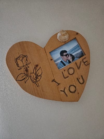 A medium-sized heart-shaped wooden plaque is affixed to a textured, light-colored wall. The plaque exhibits visible wood grain. On its left side, a pyrographically etched outline of a rose with leaves is present. The words "I LOVE YOU" are vertically etched on the right side. A rectangular photograph is embedded in the upper right portion of the plaque, showing a male wearing sunglasses being kissed on the cheek by a female. A beach and ocean are visible in the background of the photograph. A small, ribbed seashell is attached to the plaque directly above the photograph. The immediate background is an undecorated wall. No visual information within the image identifies a specific location within the Cherwell District, United Kingdom.