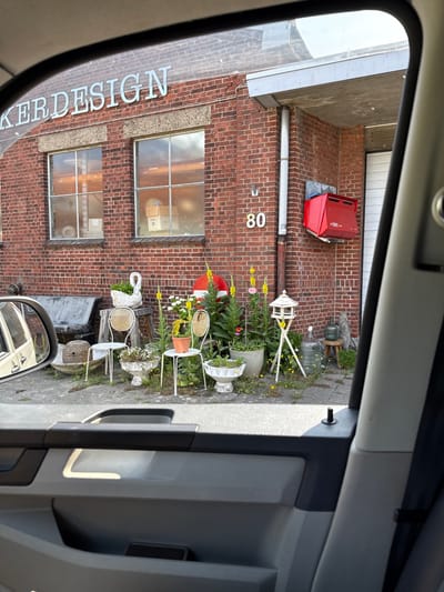 A brick building with the number 80 visible is viewed through a car window. Two windows with a visible box are present on the building, along with a red external storage unit. Decorative garden elements, including chairs repurposed as plant stands, planters, and a swan statue, line the side of the building. Part of the building sign "KERDESIGN" can be seen. A car side mirror reflecting a building is also visible.