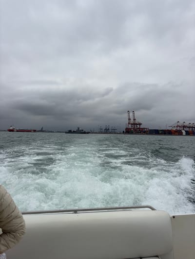 The image captures a view from a boat moving through choppy water, creating a white wake. In the distance, a cloudy sky looms over a busy industrial port. Large container cranes dominate the skyline, painted in red and white, with stacks of shipping containers visible below. Several ships are moored or navigating the waters, including a large red tanker on the left and a military or coast guard vessel to its right. The coastline features industrial structures and buildings. The perspective suggests the boat is traveling away from the shore and towards the open sea. The overall atmosphere is overcast and industrial. This scene is consistent with the Port of Callao, Peru, a major maritime hub known for its container terminals and cargo ships.