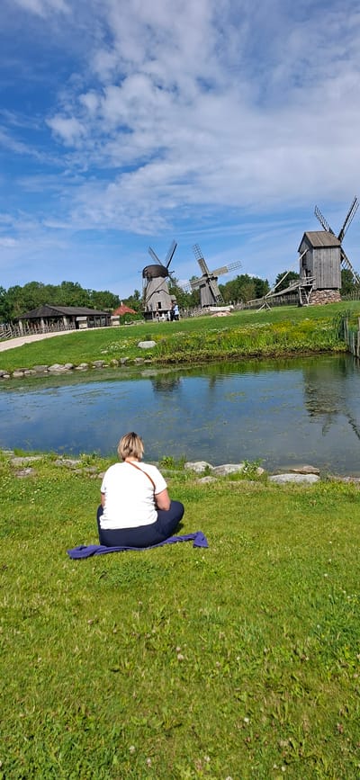 A person is sitting on a purple mat on a grassy area, facing a body of water. The person is wearing a white t-shirt and dark pants. In the background, there are several windmills. The environment is an outdoor scene with a blue sky and scattered clouds. The location is Angla, Estonia, at the Angla Heritage Windmill Park. The park features several well-preserved windmills and a pond. The person appears to be enjoying the scenery.