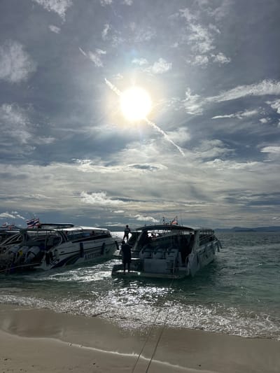 The image captures a scenic view of the water at Phru Nai, Thailand, with several speedboats docked near a sandy shore. The sun is shining brightly in a partly cloudy sky, casting a warm glow on the water. The boats, some with Thai flags visible, appear to be ready for passengers, with some individuals seen boarding or disembarking. The water is clear and calm, lapping gently against the beach. The overall impression is one of a serene coastal setting, likely a popular spot for water-based activities.