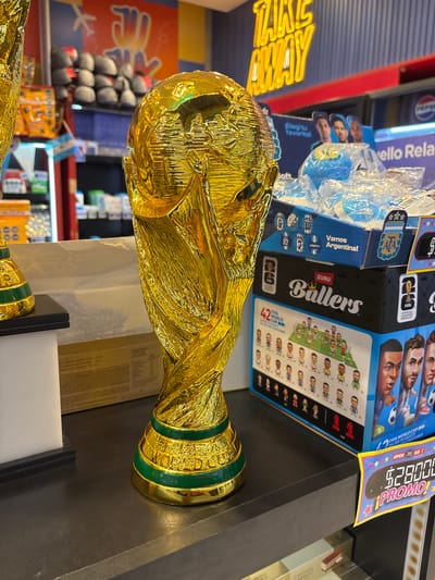 A close-up, eye-level, indoor shot features a golden replica of the FIFA World Cup trophy prominently displayed on a dark, reflective surface. The trophy, with its signature globe and sculptural figures, gleams under bright lighting. To the right of the trophy, a stack of merchandise boxes is visible, including one labeled "ZURU Ballers" featuring miniature soccer player figures and portraits of famous athletes. Another box displays the Argentine flag and the words "Vamos Argentina!" Above these boxes, a neon "TAKE AWAY" sign is partially visible against a striped blue and red background. In the background, shelves are stocked with various products, including what appear to be bags of snacks and packaged goods. A small Argentine flag is also visible to the left of the trophy. The overall environment suggests a retail setting, possibly a convenience store or souvenir shop at Aeropuerto Internacional Ezeiza, Argentina, given the presence of the World Cup trophy and Argentine-themed merchandise. The lighting is bright and artificial, creating reflections on the trophy and the counter. No people are present in the immediate foreground or interacting with the objects.