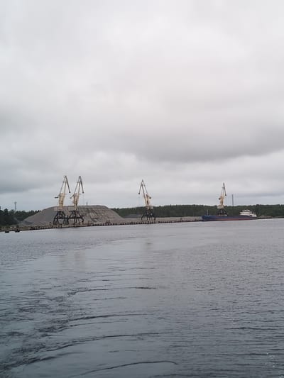 This is a wide-angle, exterior, eye-level, full shot of a cloudy day at a port facility in Shal'skii, Russia. In the foreground, choppy grey water covers most of the frame, with ripples and waves indicating movement. In the mid-ground, a stone quay runs horizontally across the image, lined with what appears to be piles of aggregate or gravel. Several large industrial cranes are positioned on the quay. On the left, two yellow and grey cranes are prominent, with one slightly in front of the other. Further to the right, another similar crane is visible. Beyond the quay, a line of green trees forms the background, with a slightly darker, dense forest further back. In the far right mid-ground, a blue and red cargo ship is docked. The sky is overcast with heavy grey clouds, obscuring the sun.