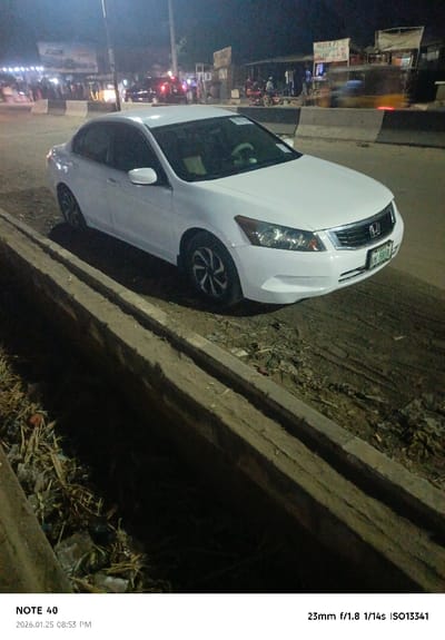 A white Honda Accord is parked on the side of a street. The car is positioned diagonally with the front facing the right. A concrete curb runs along the left side of the frame. In the background, there are buildings, streetlights, and people. Motion blur is visible, suggesting a vehicle passing. The scene is at night, with dim lighting. The license plate on the car is partially obscured but visible. Debris and trash are scattered near the curb.