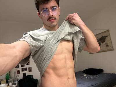 A slender man with a fair complexion gazes downward. He sports a mustache and round-framed glasses. He is partially undressed, with a gray t-shirt pulled to reveal his torso. His room features a bed with a black pillow and charger visible. A wall map hangs to his left. There are assorted bottles and photos on shelves in the background.