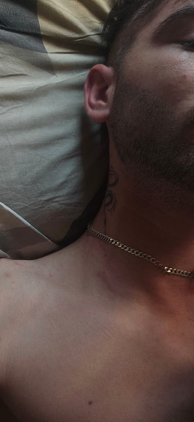 The image shows a close-up, overhead view of a person's neck and shoulder area, lying on a striped textured surface, possibly a bed or sofa. The person has dark hair, stubble on their jawline, and an ear visible. A tattoo of a coiled serpent is visible on the side of the neck, and a gold chain necklace is worn around the neck, resting on the collarbone. The skin appears slightly flushed in some areas. The overall lighting is dim and warm. There are no identifiable landmarks or elements in the image to suggest a location within Kostenets, Bulgaria.