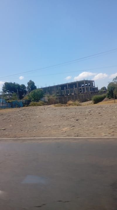 A desolate roadside view reveals a building under construction. The unfinished concrete structure stands against a clear blue sky with scattered clouds. Vegetation flanks the building, and a drab, barren landscape stretches towards the road. Power lines cross the sky above, adding to the scene's starkness. The road itself, dark and reflective, suggests recent moisture or wear.