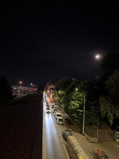 The image depicts a nighttime scene in Florence, Italy, likely from an elevated vantage point, possibly a bridge or a building overlooking a street. A road with multiple lanes of traffic is visible, with cars and motorcycles illuminated by streetlights and their own headlights. The road appears wet, reflecting the lights. On the right side of the road, there are buildings with illuminated windows and trees lining the street. On the left side, a set of train tracks or a similar railway infrastructure is visible, with several streetlights illuminating the area. The sky is dark, with a bright moon partially obscured by clouds. A few birds are seen flying in a formation in the sky. The overall impression is one of a city at night, with the moon and streetlights casting an atmospheric glow on the urban landscape.
