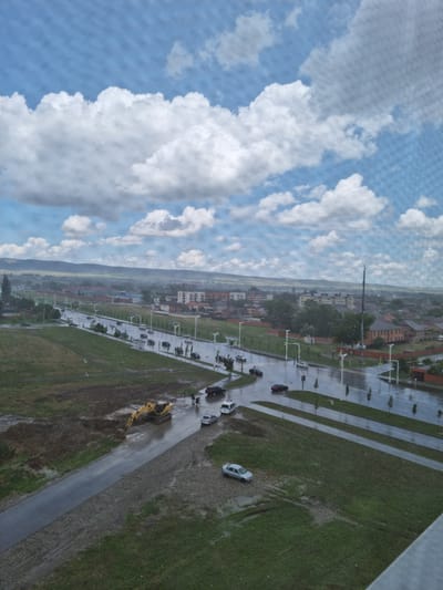 This is a high-angle, wide shot of a wet, suburban street in Grozny, Russia, under a partly cloudy sky. The road, slick with rain, is lined with streetlights and trees. Several cars are driving on the road, and a yellow excavator is parked on the side, next to a construction site. In the background, there are residential buildings and a distant mountain range. The overall impression is of a damp, overcast day in a developing area.