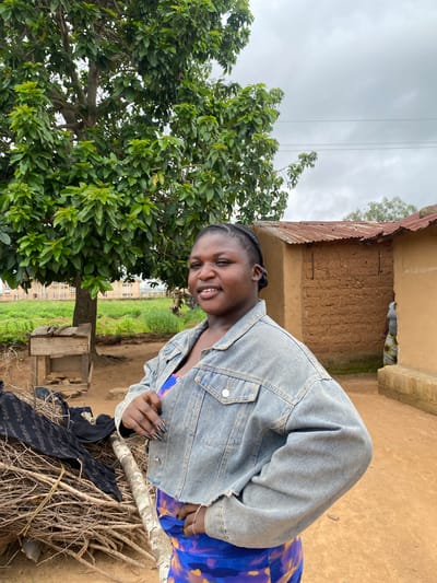 A young woman with braided hair and dark nail polish stands outdoors, posing for the camera with one hand on her hip. She is wearing a light blue denim jacket over a colorful patterned dress. Behind her, a large tree with green leaves stands to the left, and to the right are two simple, mud-brick structures with corrugated metal roofs. The ground is made of packed dirt. In the background, there is lush green vegetation and a cloudy sky. The scene appears to be in a rural area of Riyom, Nigeria.