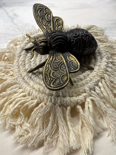 A high-angle, close-up shot depicts a metal bee figurine resting on a circular, fringed coaster. The bee is dark, possibly black, with gold-colored decorative wings. The coaster is made of a light-colored, possibly off-white, material with a thick, knotted center and long, loose fringe extending outward. The surface beneath the coaster appears to be a light-colored, possibly white, textured surface. The image does not include details that would confirm its location as Jimbaran, Indonesia.