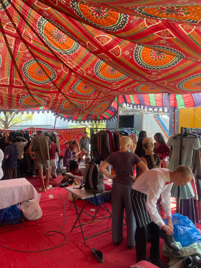 The image depicts the interior of a tent-like structure, possibly a marketplace in Anajpur, India, judging by the textiles. The tent is covered with red fabric featuring orange and blue floral patterns. Several people, dressed in varying attires, are browsing clothing hung on racks. One man, wearing a white shirt and a tie, is trying on a black shoe. Other individuals, including women with braided hair and men in casual clothing, are seen looking at the merchandise. The floor is covered in red fabric, and electrical cords snake across it. A rack and an electrical outlet are visible.