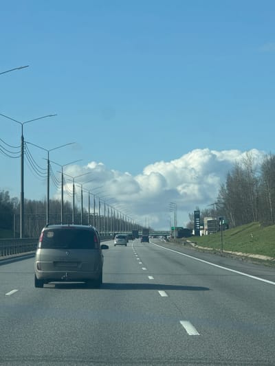 A landscape photograph depicts a multi-lane highway under a clear blue sky with cumulus clouds. A gray minivan occupies the right-hand lane, followed by other vehicles in the distance. Parallel to the road, a series of utility poles extend towards the horizon, their wires visible against the sky and clouds. A grassy embankment and trees border the highway on the right. In the middle distance, a gas station sign can be identified. The orientation of the image is sideways, requiring rotation for correct viewing. There is no information available to confirm it is in the city of Nikol'skoe, Russia.