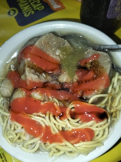 The image displays a white, shallow bowl containing an Indonesian dish, likely bakso, situated on a vibrant yellow surface in Padangkleng, Indonesia. The bowl's contents include a base of pale yellow thin noodles, several irregularly shaped greyish-brown meat pieces (suggesting meatballs or sliced meat), and a translucent, light-colored broth. The dish is heavily adorned with a viscous, reddish-orange sauce applied in broad drizzles across the meat and noodles. Streaks of a darker, possibly sweet soy sauce, are mixed within the red sauce. A lighter, translucent greenish-yellow condiment is also visible on the central meat piece. A metallic spoon is partially submerged within the broth on the right side of the bowl. The yellow surface beneath the bowl features partially visible black and blue text reading "KECAP MANIS" and "food" in the upper left quadrant. A dark-colored bottle with a faint label is present in the upper right background. No individuals are visible.