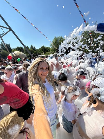 The image shows a sunny outdoor event, likely a foam party, in the city of Zavolzhye, Russia. In the foreground, a woman with long, blonde, braided hair smiles at the camera, her arm outstretched as if taking a selfie. She is wearing a white tank top and denim shorts. She is surrounded by a crowd of people, many of whom are children, all covered in white foam. The foam is being sprayed from a large blue machine visible in the upper right portion of the image. In the background, there are trees, a dome-shaped structure, and colorful triangular flags strung across the sky. Some individuals are holding red and green balloons. The overall atmosphere appears to be festive and playful.