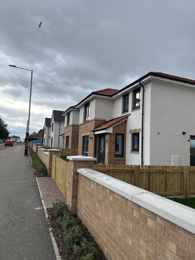 The image shows a street scene in Dunfermline, United Kingdom, with a row of modern houses along one side. The houses are two-story, with a mix of brick and white rendered facades, and dark-framed windows. Each house has a small front garden enclosed by a low brick wall topped with a white coping stone and wooden fences.
A paved sidewalk runs along the front of the houses, with a narrow strip of vegetation between the sidewalk and the road. The road is asphalt and appears wet. A street light is visible on the left, extending into the cloudy sky. In the distance, cars are parked along the street. A bird flies in the sky.