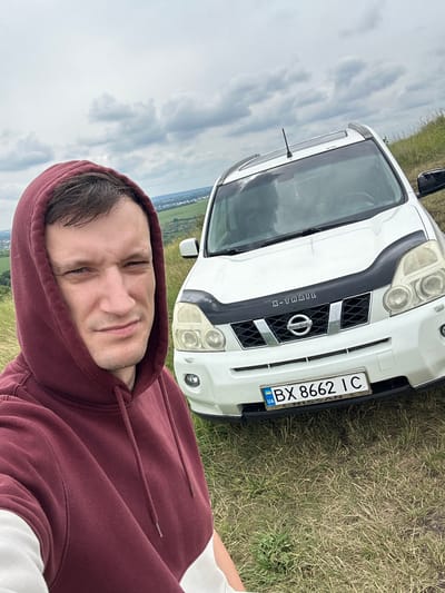 In Lysohirka, Ukraine, a man wearing a maroon hooded sweatshirt stands in front of a white Nissan X-Trail SUV. The man is facing the camera and appears to be taking a selfie, with the car and a cloudy sky in the background. The car has a license plate that reads "BX 8662 IC". The scene is set in a grassy, outdoor environment, possibly a hilltop, with vegetation visible.