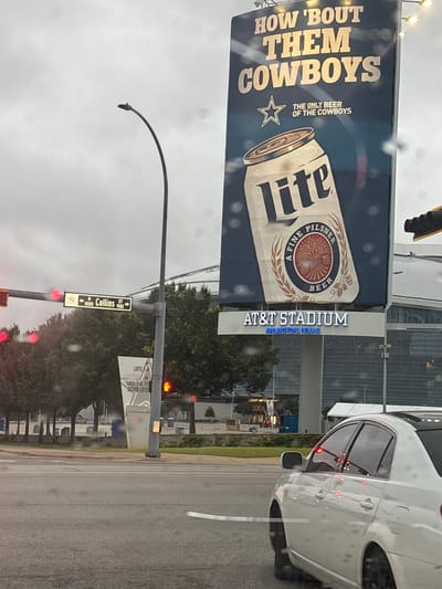 The image shows a street scene in Arlington, Texas, with AT&T Stadium in the background, home of the Dallas Cowboys NFL team. Prominently displayed is a large billboard advertising Miller Lite beer with the slogan "HOW 'BOUT THEM COWBOYS". Street signs indicate "N Collins St" for the 1000 and 900 blocks. A white car is partially visible on the right side of the frame. The weather appears overcast, with raindrops on the windshield. Additional signage points to stadium parking for lots 7-9, 11, and Silver Lots. A traffic light is visible, indicating the presence of an intersection.