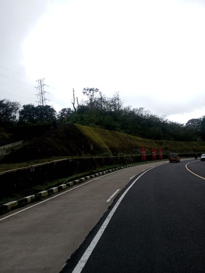This image depicts a roadside scene within the city of Deothang, Bhutan. A multi-lane asphalt road, marked with white and yellow lines, curves right, receding from the foreground. To its left, a lighter gray paved shoulder is delineated by a white line and a curb with black and white stripes. Beyond the curb, a dark concrete retaining wall supports a steep, multi-tiered earthen embankment covered predominantly with dark soil and patches of green moss/vegetation. Dense green trees and brush crown the embankment and extend into the background.
Two human figures are visible on the upper slope of the embankment, near the tree line. One figure, dressed in dark clothing, stands on the left. A second figure, lighter in color, is positioned further right. A large metal lattice transmission tower, carrying power lines, stands on the upper left portion of the embankment. Three red vertical banners, each displaying white text (partially legible as "FLOW" and "BHUTAN") and a right-pointing white arrow, are affixed along the lower retaining wall of the embankment. On the main road, a light-colored van is observed in the mid-ground, followed by a motorbike and a lighter-colored car, all moving away from the camera. The sky is overcast and uniformly light gray.