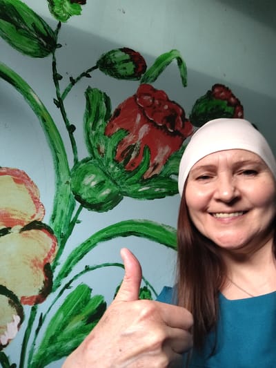 A smiling woman is captured in a close-up, chest-up shot, giving a "thumbs up" gesture. She is positioned on the right side of the frame, facing slightly left with a broad, genuine smile that shows her teeth. Her medium-length brown hair frames her face, and she wears a light-colored (likely white or off-white) headband or thin cap. Her attire includes a teal or blue-green top. The woman's expression conveys happiness and approval.
Behind her is a wall adorned with a vibrant, hand-painted floral mural. The wall itself is painted a light blue or teal color, serving as a backdrop for large, stylized flowers and foliage. The flowers feature prominent red and yellow/cream petals with dark outlines, while the leaves and stems are depicted in various shades of green, characterized by visible brushstrokes, giving the mural an artistic, perhaps folk-art, aesthetic. The lighting is bright and appears even, suggesting an indoor setting or a softly lit environment. There is no visible text in the image. The scene takes place in Сусады-Эбалак, Russia.