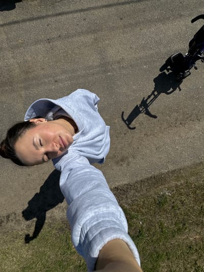 A woman in a gray hoodie lies on a paved surface near a grassy area, taking a selfie with her arm extended. A bicycle and its shadow are visible in the upper left corner of the frame. The scene appears to be outdoors on a sunny day. Location cannot be determined.