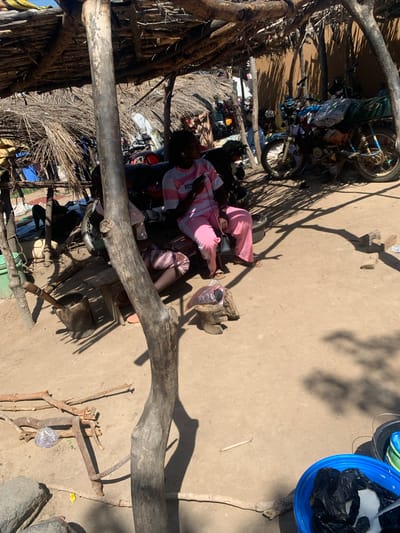 This media file shows an outdoor scene in Dull A, Nigeria. The image is oriented vertically. In the foreground, a large, weathered tree branch stretches horizontally across the frame, casting a shadow. To the left, a rustic structure made of branches and dried vegetation suggests a simple dwelling or market stall. Inside this structure, several motorbikes are visible, some with items piled on them.

Further into the scene, two individuals are seated on the ground. One person, wearing a pink shirt and dark pants, is leaning back. The other individual, also seated, wears a pink and white striped garment. They appear to be resting or engaged in quiet activity. Scattered around them are various objects, including plastic bags, what looks like a basket, and wooden items.

In the background, the ground is dusty and uneven. The bright sunlight indicates it is daytime, and the strong shadows suggest a sunny weather condition. To the right of the frame, a blue plastic bucket contains black plastic bags, and there are other buckets or containers nearby. The overall impression is of a rural or semi-urban setting in Nigeria, with a focus on everyday life and the presence of motorbikes, which are common modes of transportation. The scene appears peaceful, with the individuals resting in the shade. No visible text is discernable in the image.