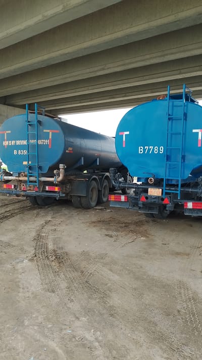 The image shows two blue tanker trucks parked in Effurun, Nigeria. The trucks are positioned beneath a concrete overpass. The ground is dirt with tire tracks. The first truck displays the text "HOW IS MY DRIVING 09032067311" and "B 8359." The second truck displays "B 7789" on its tank. Each truck has a ladder leading to the top of the tank and a red and white "T" logo.