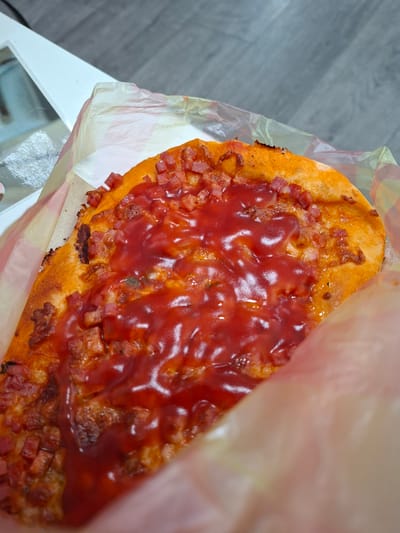 A partially eaten pizza slice is presented inside a partially open plastic takeout bag on a white table with a reflective surface. The pizza features a red sauce covering a topping of small, cubed pieces of a cured meat product. The crust appears to be slightly burnt along the edge. The table sits on a gray wood-patterned floor. It is unclear if this is Galabovo, Bulgaria.