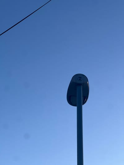 The image shows a streetlight fixture against a clear blue sky. The fixture is dark in color and mounted on a horizontal pole. In the lower left corner, a thin black wire stretches across the frame. The location is Tromsø, Norway, but that is not explicitly visible.