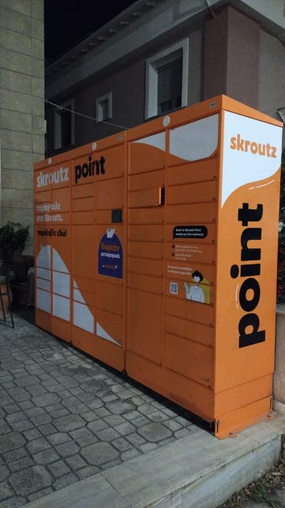 This media file captures an orange parcel locker, branded with "skroutz point," situated outdoors. The locker is a self-service delivery point, likely in Greece given the Greek text on its surface. The prominent text on the top reads "point" in large black lettering, and "skroutz" in orange to its left. The locker itself is divided into multiple compartments, suggesting it's designed for package retrieval and drop-off.
Below the "point" text, there's an informational panel with instructions in Greek. One section explains that the locker can be opened in two ways: via a code received on a mobile phone or through Bluetooth from the Skroutz app. Another blue panel highlights "δωρεάν μεταφορικά," meaning "free shipping." Further down, more Greek text appears, reading "παράγγειλε στο Skroutz, παράλαβε εδώ!", which translates to "order from Skroutz, receive here!".
The setting appears to be a street or sidewalk, with cobblestone paving visible in the foreground and a textured wall or building in the background. The lighting suggests it might be evening or nighttime, as the surroundings are somewhat dimly lit, although the locker itself is well-illuminated. The overall impression is of a functional, modern piece of urban infrastructure facilitating online shopping logistics.