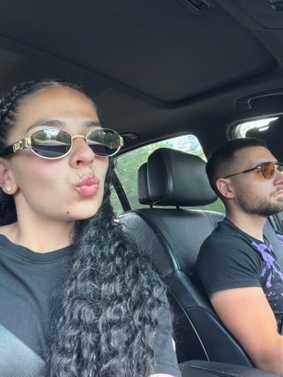 This media file shows two people, a man and a woman, in the interior of a car. The woman is in the foreground, positioned closer to the camera and appears to be taking the selfie. She has dark, curly hair and is wearing large, oval-shaped sunglasses with gold frames. Her lips are pursed. She is wearing a black t-shirt.
The man is in the background, sitting in the passenger seat. He is wearing sunglasses with an octagonal amber lens and a black t-shirt with a purple graphic design. He is looking upwards and to the right, away from the camera. His arm is resting on the seat.
The scene appears to be outdoors, indicated by the greenery visible through the car windows. The lighting suggests it is daytime. The overall mood seems casual and relaxed, possibly a road trip. There is no visible text in the image. The location context is Brusovete, Bulgaria.