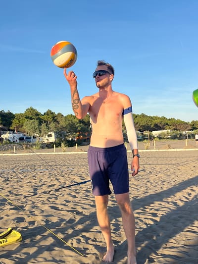 The image depicts a man spinning a volleyball on his finger on a sandy beach in Pistula, Montenegro. He is wearing dark blue shorts, sunglasses, and a white arm sleeve. The background shows trees, parked vehicles, and a clear blue sky. The lighting suggests it is late afternoon or early evening.