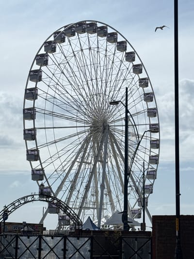 The image captures a large Ferris wheel, dominating the frame against a bright, overcast sky. Its intricate white structure of spokes and numerous grey cabins, some with subtle purple window tints, is clearly visible. No people are present on the ride or in the surrounding scene.
A bird is captured in mid-flight in the upper left quadrant of the sky, adding a natural element to the urban-like landscape.
On the right side of the image, dark architectural elements frame the Ferris wheel. These include a substantial metal framework with a grid pattern, suggesting part of an adjacent building or another amusement park attraction, and a section of a brick wall visible above it. A yellow sticker bearing the black numbers "23693" is affixed to the brickwork. A dark lamppost or utility pole extends vertically into the frame near the center-right, casting a shadow across a portion of the Ferris wheel's structure. In the lower right, an arched, decorative metal structure, possibly a smaller ride or an entrance archway, features some illegible graffiti and a partially visible blue and white flag or banner.
The scene depicts an outdoor leisure or amusement area, consistent with the coastal setting of Gorleston-on-Sea, United Kingdom. It is daytime, under a bright yet cloudy sky, suggesting fair weather. The image conveys a sense of an attraction ready for activity, though currently stationary.
