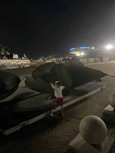 A young boy wearing a white t-shirt and red shorts stands on a scooter next to a large, dark, metallic object, possibly an anchor, at night. The boy is facing away from the camera and appears to be touching the object. The scene is illuminated by artificial lights, casting long shadows. In the background, a large ship is docked, and a building with illuminated blue lettering, reading "NOVOROSSIYSK," is visible. The ground is paved with brick-like stones. The overall environment suggests a port or waterfront area in the city of Novorossiysk, Russia.