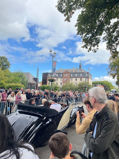 This image depicts a public event occurring outdoors on a street in Molsheim, France. A two-tone, black and cream/tan vintage convertible car, with its soft top folded down, occupies the midground, moving left to right. Two individuals are visible in the car's rear seating area, one in a white shirt and cap. Spectators line both sides of the street, separated by metal barriers, observing the passing vehicle. Several individuals in the foreground, including a male with grey hair and glasses, and another person in a tan hoodie, are actively recording the event with smartphones. In the background, a prominent multi-story building featuring red brick walls, a grey roof, and a central spire is visible, identified as the Hôtel de la Monnaie, a landmark in Molsheim. A streetlight pole to the left of this building displays a blue banner with the white "Bugatti" logo. The sky is partially cloudy with patches of blue, and green foliage from trees frames the scene on both sides.