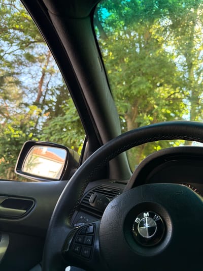 The image shows the interior of a car, looking out through the windshield and side window. The steering wheel is visible in the foreground, with the BMW logo prominently displayed on the center. To the left, a side mirror reflects a building with a red-tiled roof. Outside the car, lush green trees are seen, with sunlight filtering through the leaves, creating a bokeh effect. The scene suggests a sunny day, possibly in a suburban or rural setting, as indicated by the presence of trees and a house. There are no people visible in the image. The car appears to be parked or moving slowly. The location is identified as Dinkata, Bulgaria.