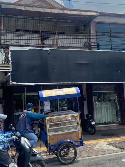 The image shows a street scene in Mojokerto, Indonesia, at the coordinates -7.467 latitude and 112.44 longitude. A man wearing a blue vest, a white shirt, and a blue cap is sitting on a motorcycle with a blue and yellow food cart attached. The cart is loaded with various food items, visible through a glass display. The man appears to be in the process of driving the cart, as he is looking ahead and his right hand is on the handlebar. Behind him, a storefront with a large black sign is visible. The sign reads "Setenganpra Oloh-oleh". In the storefront window, white garments can be seen. A motorcycle is parked in front of the store. The scene is set on a paved street with yellow painted lines. The sidewalk is made of grey bricks and a yellow painted curb borders the road. The weather is sunny.