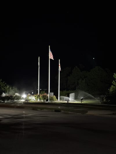 The image shows the Panama City Beach Veterans Memorial at night. Three flags are prominently displayed on tall poles. The leftmost flag is white with a red cross. The middle flag is the American flag. The rightmost flag is white with a red cross and a white field in the canton. The memorial features a white monument or plaque with text that is partially legible, reading "PANAMA CITY BEACH VETERANS MEMORIAL". In front of the memorial, a park bench is visible, and sprinklers are watering the surrounding green area. The scene is illuminated by artificial lights, creating a stark contrast with the dark night sky. The ground in the foreground appears wet, possibly from rain or the sprinklers. Trees and bushes form the background.