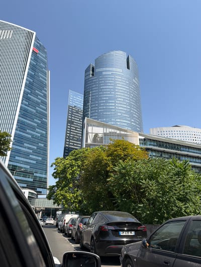 The image depicts a street scene in Nanterre, France, characterized by modern architecture and moderate traffic. In the foreground, a car's interior is visible from the driver's perspective, looking out towards the street. The street is lined with several vehicles, including a black Tesla with license plate "GH 438 HK," indicating it is a recent model electric car. The vehicles are stationary or moving slowly, suggesting traffic congestion.
The background is dominated by several large, contemporary buildings, typical of a business district. To the left, a tall, slender skyscraper with a white and blue facade stands prominently. To the right and center, a large, curved skyscraper with a glass facade reflects the clear blue sky. Another, narrower skyscraper with a dark glass facade is visible between these two structures. Lower buildings with white facades and numerous windows are also present, some with terraced levels covered in greenery. Lush green trees line the street and are interspersed between the buildings, providing a contrast to the urban landscape. The sky is a vibrant, cloudless blue, suggesting a sunny day. The overall impression is one of a modern, urban environment with a mix of business infrastructure and natural elements.