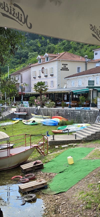 The image depicts a waterfront scene in Rijeka Crnojevića, Montenegro. In the foreground, a white and red boat is partially submerged in the water, with wooden pallets and tires scattered along the shoreline. Adjacent to the water, a vibrant green carpet covers an area of grass, with a yellow plastic container placed on it. Further back, a collection of colorful kayaks and canoes are neatly arranged on the lawn, suggesting a rental or recreational area.
Behind the kayaks, a stone wall separates the grassy area from a paved walkway leading up a series of concrete steps. Beyond the steps, a row of buildings lines the waterfront. One building, a hotel, is prominently featured with a sign reading "HOTEL S FAMILY JOVICEVIC." Another building appears to be a restaurant with outdoor seating. The architecture is consistent with traditional coastal European buildings, featuring tiled roofs and light-colored facades.
Overhead, a large white awning partially obscures the top of the image, with text visible in a stylized script. The background is dominated by lush green trees and hills, indicating a mountainous or heavily forested environment surrounding the town. The overall impression is one of a peaceful, picturesque location, likely a tourist destination with amenities for water activities and dining. No people are clearly visible in the image, but the presence of outdoor seating at the restaurant implies potential patrons. The scene is bathed in natural daylight, suggesting it was taken during the day.
